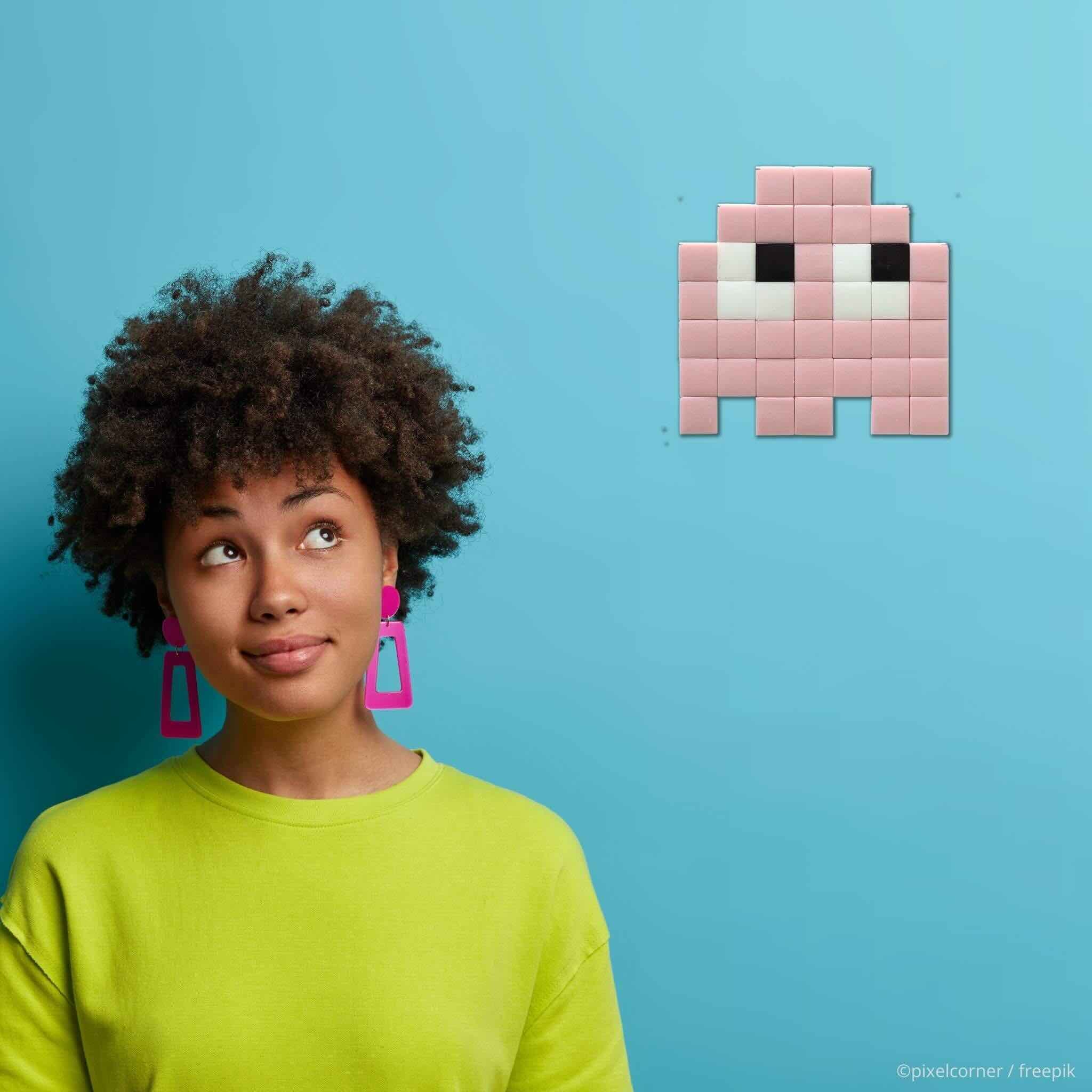Une femme jeune sur fond bleu regarde du coin de l'oeil un fantome rose en pixel art simple collé sur un mur en décoration, le coffret de mosaïque diy Les Gloomie(s)
